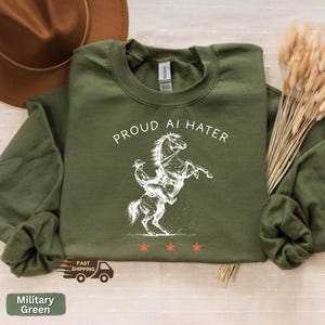 May include: Military green sweatshirt with the text "PROUD AI HATER" above a white illustration of a horse rearing up with a rider. Three red stars are below the illustration. The sweatshirt has long sleeves.