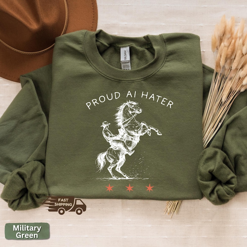 May include: Military green sweatshirt with the text "PROUD AI HATER" above a white illustration of a horse rearing up with a rider. Three red stars are below the illustration. The sweatshirt has long sleeves.