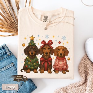 May include: Ivory-colored t-shirt featuring three dachshunds. One dog is dressed as a Christmas tree, another with a red bow, and the third in a red sweater. The shirt has a Comfort Colors tag.