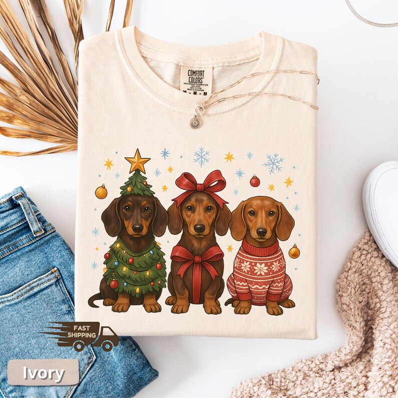 May include: Ivory-colored t-shirt featuring three dachshunds. One dog is dressed as a Christmas tree, another with a red bow, and the third in a red sweater. The shirt has a Comfort Colors tag.