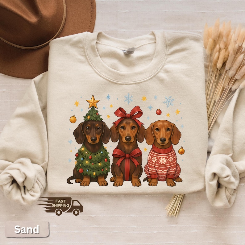 May include: A cream-colored sweatshirt featuring three dachshunds in festive holiday attire. One dog is adorned with a Christmas tree, another with a large red bow, and the third in a red sweater. The design includes snowflakes and star accents.