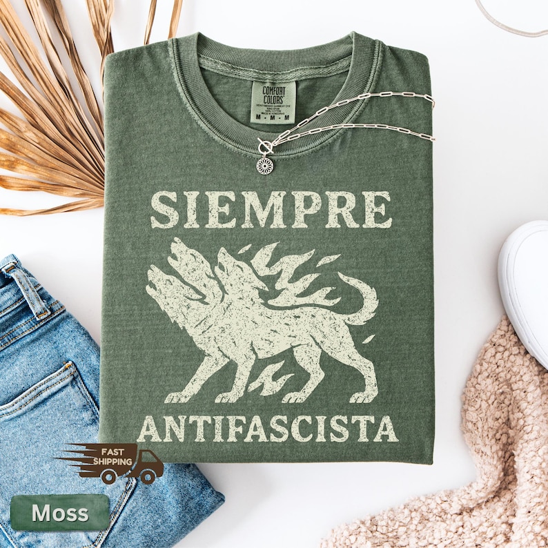 May include: Moss green t-shirt with the words "SIEMPRE ANTIFASCISTA" in white block letters. A three-headed dog graphic is centered below the text. A silver necklace with a sun pendant rests on the shirt.