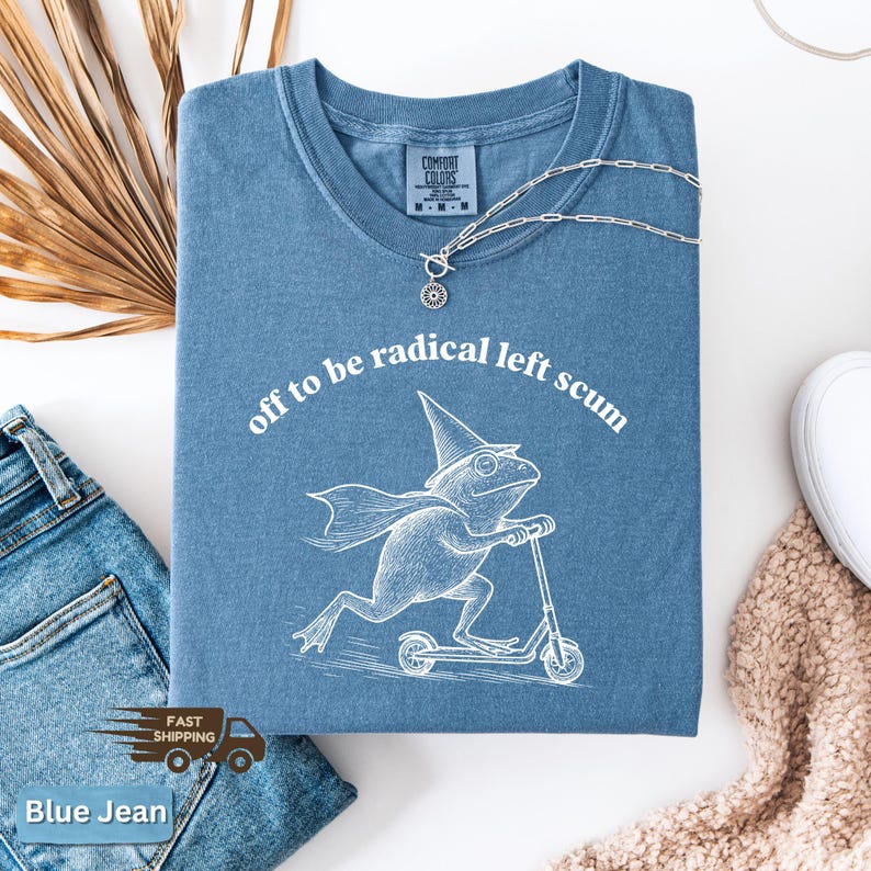 Pode incluir: Uma camiseta azul com a frase "off to be radical left scum" e um gr&aacute;fico de um sapo numa scooter. Um colar de prata com um pingente est&aacute; sobre a camiseta. Jeans azuis e um su&eacute;ter bege no fundo.