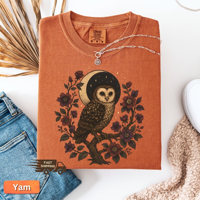 May include: A rust-coloured Comfort Colors t-shirt with an owl design, surrounded by flowers and a crescent moon. A silver necklace with a sun pendant is on the shirt. The t-shirt has a crew neck and short sleeves.