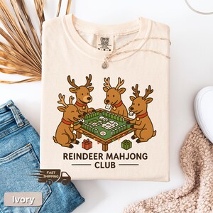 Puede incluir: Camiseta de color marfil con una caricatura de cuatro renos jugando al mahjong alrededor de una mesa verde. El texto "REINDEER MAHJONG CLUB" est&aacute; impreso debajo. La camiseta est&aacute; doblada, con un collar y unos vaqueros visibles.