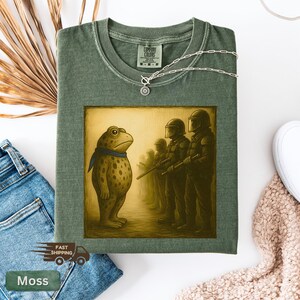 K&ouml;nnte beinhalten: Moosgr&uuml;nes T-Shirt mit einer Grafik einer Froschfigur, die Soldaten gegen&uuml;bersteht. Der Frosch tr&auml;gt einen blauen Schal. Das Shirt hat ein Comfort Colors-Etikett und eine silberne Halskette. Die Worte "Fast Shipping" sind sichtbar.