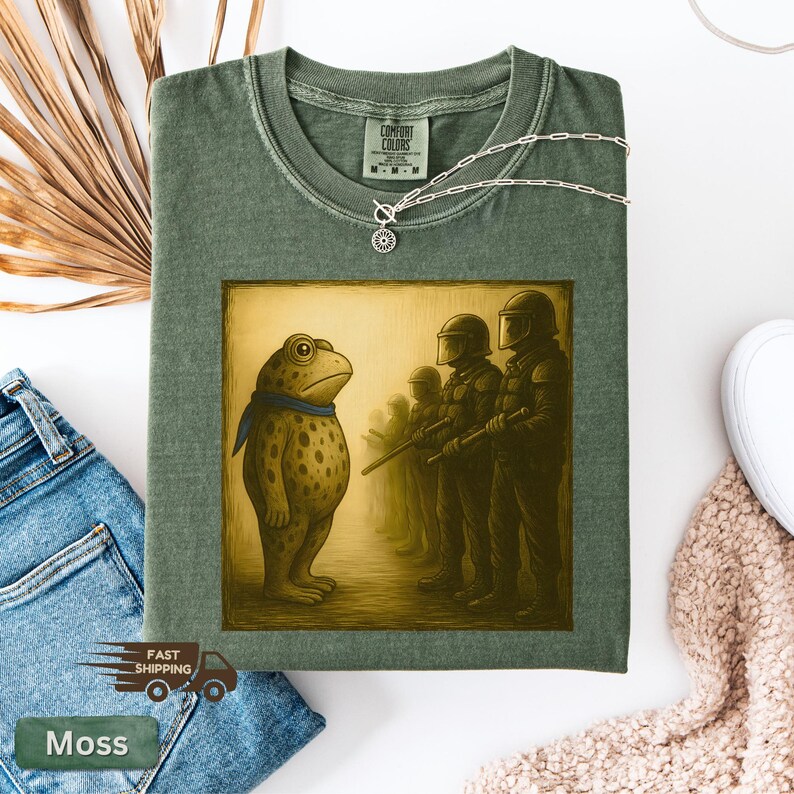 K&ouml;nnte beinhalten: Moosgr&uuml;nes T-Shirt mit einer Grafik einer Froschfigur, die Soldaten gegen&uuml;bersteht. Der Frosch tr&auml;gt einen blauen Schal. Das Shirt hat ein Comfort Colors-Etikett und eine silberne Halskette. Die Worte "Fast Shipping" sind sichtbar.