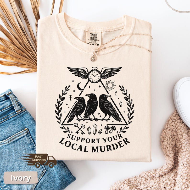 May include: Ivory-colored t-shirt with a black graphic featuring three crows inside a triangle, a clock, and the text "SUPPORT YOUR LOCAL MURDER." The shirt has a crew neck and short sleeves.