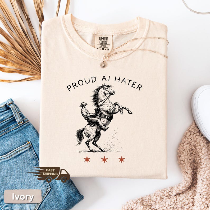 May include: Ivory-colored t-shirt with the text "PROUD AI HATER" above a black and white illustration of a cowboy riding a rearing horse. Three red stars are below the illustration. The shirt is made of a soft material.