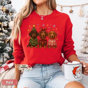 May include: Red sweatshirt featuring three dachshunds in festive holiday attire. One dog is dressed as a Christmas tree, another with a bow, and the third in a sweater. Snowflakes and stars decorate the background.