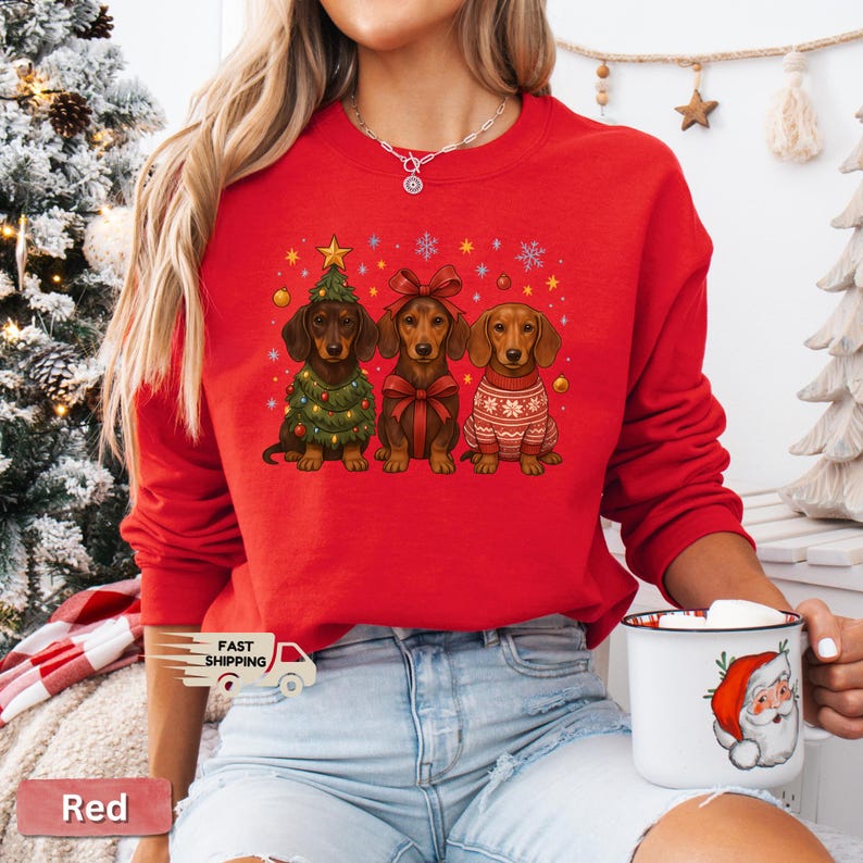 May include: Red sweatshirt featuring three dachshunds in festive holiday attire. One dog is dressed as a Christmas tree, another with a bow, and the third in a sweater. Snowflakes and stars decorate the background.