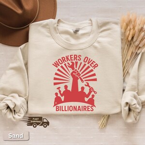 May include: Sand-colored sweatshirt with a red graphic that reads "WORKERS OVER BILLIONAIRES." The design features a raised fist holding tools, with silhouettes of workers below. The sweatshirt has long sleeves.