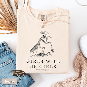 May include: Ivory-colored t-shirt with a black graphic of a praying mantis wearing a crown, with the text "GIRLS WILL BE GIRLS" and "Mantis religiosa" below. The shirt is folded and displayed with a pair of blue jeans and a necklace.