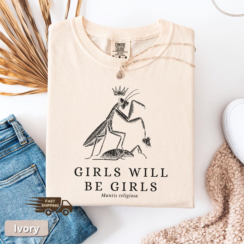 May include: Ivory-colored t-shirt with a black graphic of a praying mantis wearing a crown, with the text "GIRLS WILL BE GIRLS" and "Mantis religiosa" below. The shirt is folded and displayed with a pair of blue jeans and a necklace.