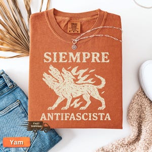 May include: Burnt orange t-shirt with the words "SIEMPRE ANTIFASCISTA" and a three-headed wolf graphic. The shirt is folded and has a Comfort Colors tag. A silver necklace with a sunburst pendant rests on the shirt.