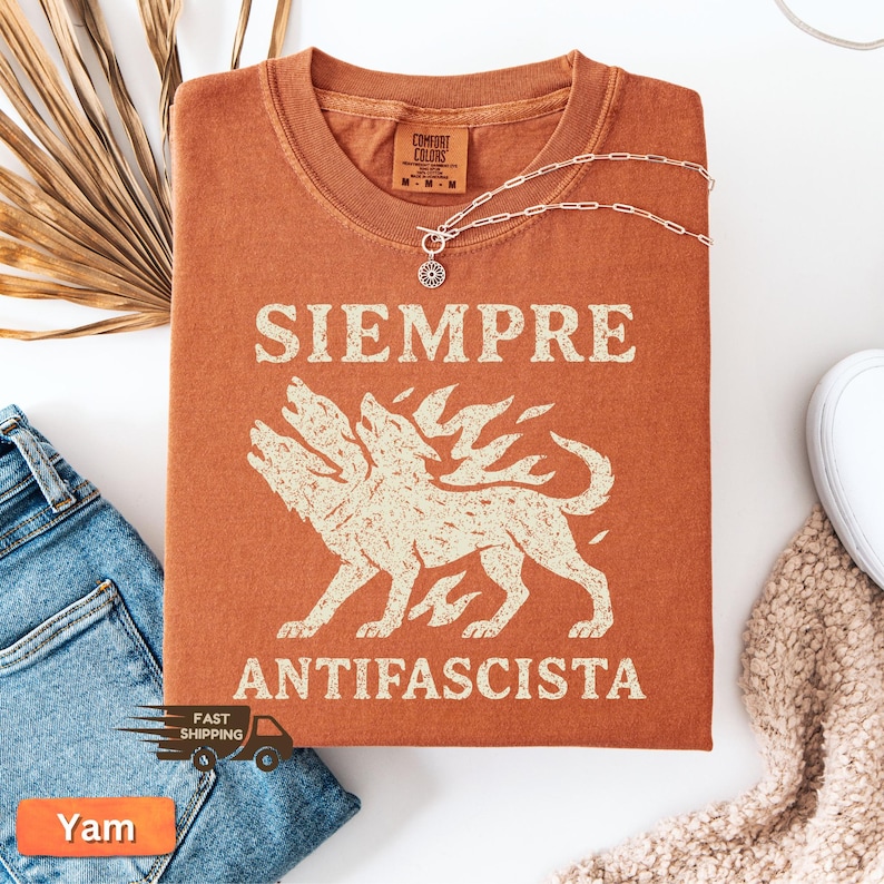 May include: Burnt orange t-shirt with the words "SIEMPRE ANTIFASCISTA" and a three-headed wolf graphic. The shirt is folded and has a Comfort Colors tag. A silver necklace with a sunburst pendant rests on the shirt.