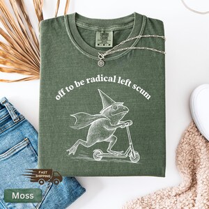 Pode incluir: Camiseta verde musgo com a frase "off to be radical left scum" em arco. Uma ilustra&ccedil;&atilde;o branca de um sapo com chap&eacute;u de mago e capa em uma scooter est&aacute; abaixo do texto. Um colar de prata est&aacute; sobre a camiseta.