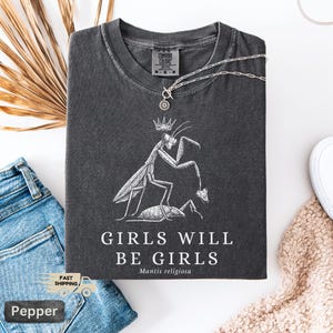 May include: Dark gray t-shirt with a white graphic of a praying mantis wearing a crown, with the text "GIRLS WILL BE GIRLS" and "Mantis religiosa". A silver necklace with a pendant is on the shirt. The shirt is folded on a white surface.
