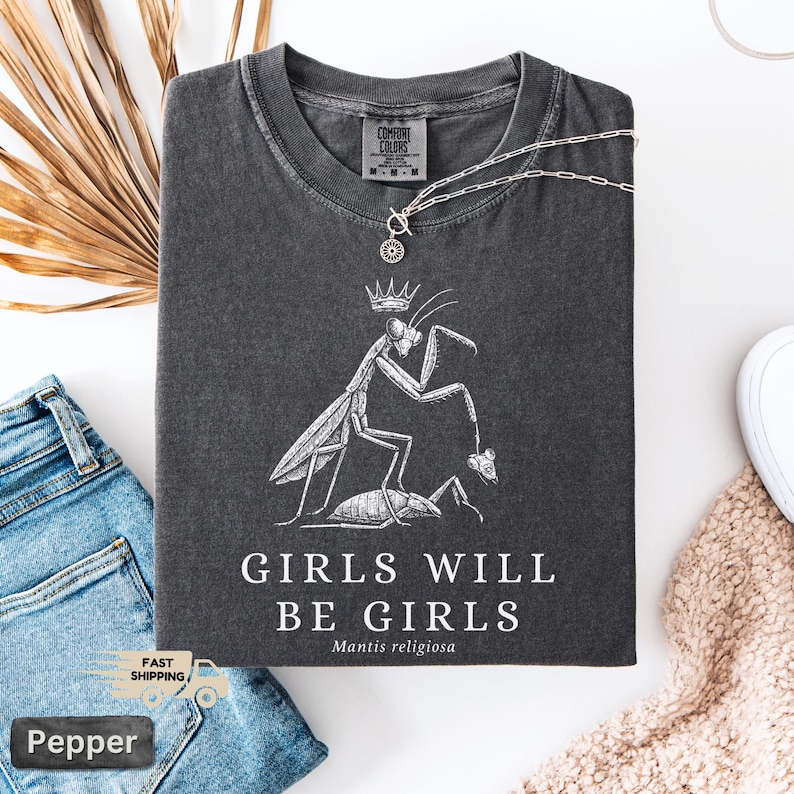 May include: Dark gray t-shirt with a white graphic of a praying mantis wearing a crown, with the text "GIRLS WILL BE GIRLS" and "Mantis religiosa". A silver necklace with a pendant is on the shirt. The shirt is folded on a white surface.