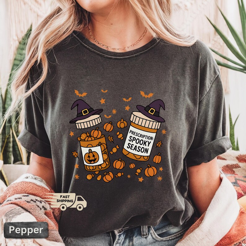 Op de afbeelding: Donkergrijs t-shirt met een Halloween-thema. De afbeelding toont twee pillenflesjes met heksenhoeden, gevuld met pompoenen en de tekst "Prescription: Spooky Season". Oranje pompoenen en vleermuizen zijn verspreid rond de flesjes.