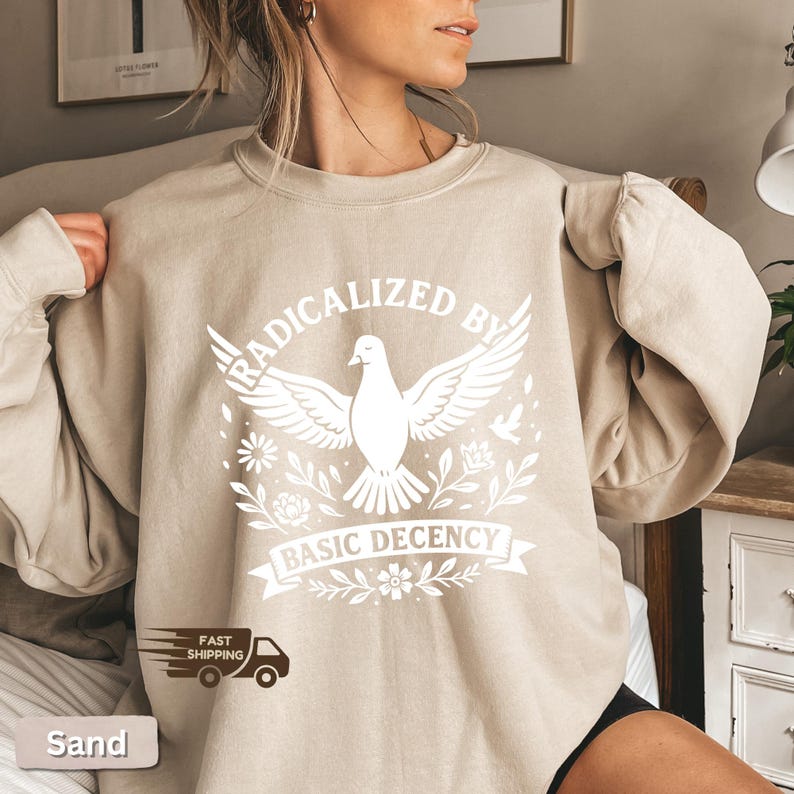Op de afbeelding: Zandkleurige sweatshirt met een witte afbeelding van een duif met uitgespreide vleugels, omringd door bloemmotieven. De tekst "RADICALIZED BY BASIC DECENCY" staat boven en onder de duif gedrukt. De sweatshirt heeft lange mouwen.