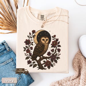 May include: Ivory t-shirt with an owl illustration. The design features a brown and white owl on a branch, encircled by burgundy flowers and a crescent moon against a starry sky. The shirt has the text "Comfort Colors".