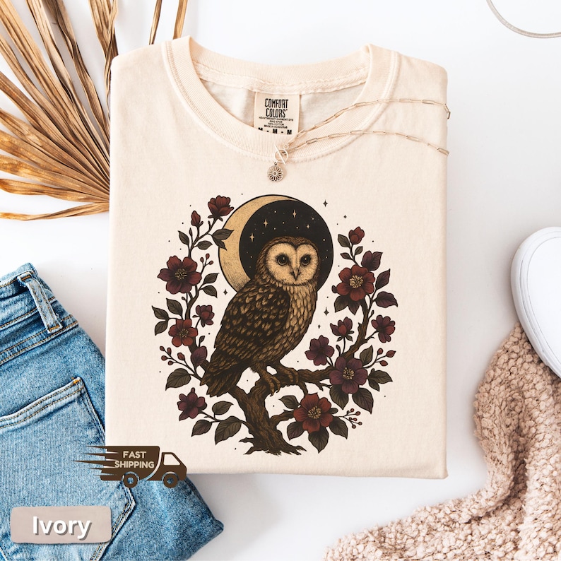 May include: Ivory t-shirt with an owl illustration. The design features a brown and white owl on a branch, encircled by burgundy flowers and a crescent moon against a starry sky. The shirt has the text "Comfort Colors".
