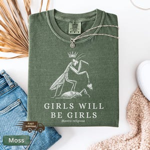 May include: Moss green t-shirt with a white graphic of a praying mantis wearing a crown and holding a smaller insect. The text "GIRLS WILL BE GIRLS" is below the graphic. A silver necklace is draped across the shirt.