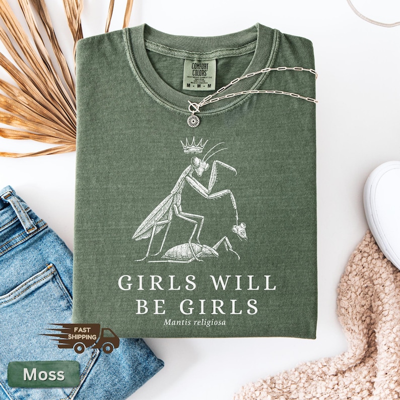 May include: Moss green t-shirt with a white graphic of a praying mantis wearing a crown and holding a smaller insect. The text "GIRLS WILL BE GIRLS" is below the graphic. A silver necklace is draped across the shirt.