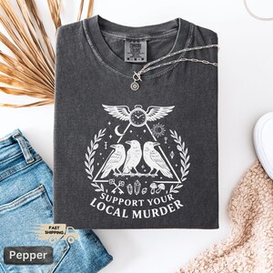 May include: Dark gray Comfort Colors t-shirt with a white graphic featuring three crows, a clock, and the text "SUPPORT YOUR LOCAL MURDER." A silver necklace with a pendant rests on the shirt. The shirt is folded on a white surface with blue jeans.