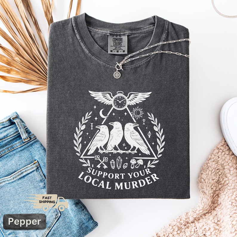 May include: Dark gray Comfort Colors t-shirt with a white graphic featuring three crows, a clock, and the text "SUPPORT YOUR LOCAL MURDER." A silver necklace with a pendant rests on the shirt. The shirt is folded on a white surface with blue jeans.