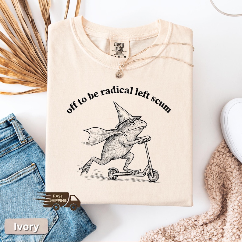 Pode incluir: Camiseta cor marfim com a frase "off to be radical left scum" acima de um gr&aacute;fico de um sapo andando de scooter, usando um chap&eacute;u de mago e capa. A camiseta &eacute; feita de um material macio.
