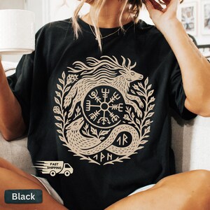以下が含まれることがあります： 黒いTシャツ。ベージュの円形デザインで、神話上の生き物とルーン文字が描かれています。デザインは葉やその他の装飾要素で囲まれています。ゆったりとしたフィット感のシャツです。