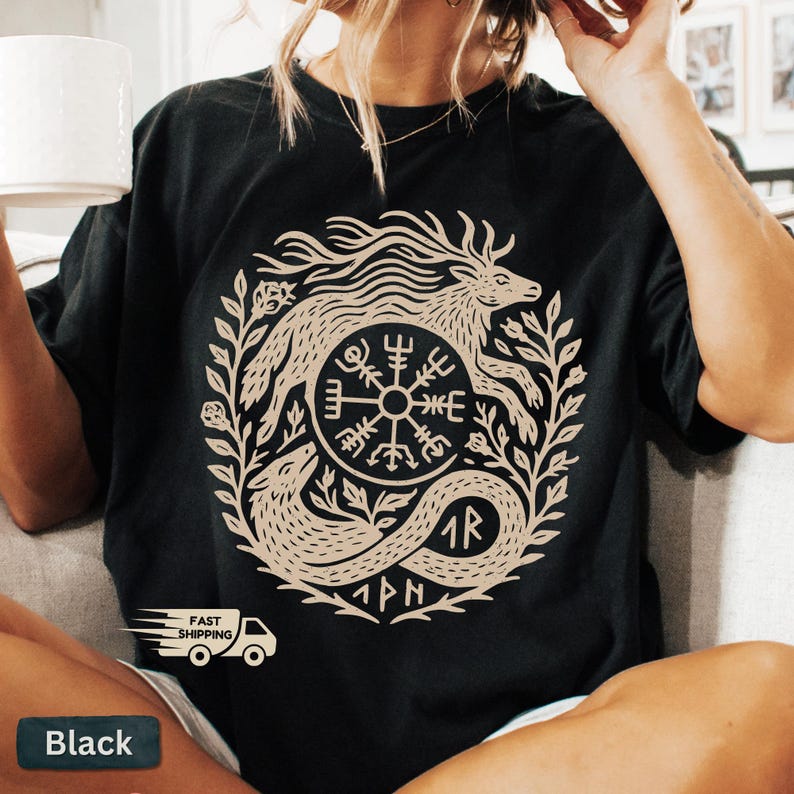 以下が含まれることがあります： 黒いTシャツ。ベージュの円形デザインで、神話上の生き物とルーン文字が描かれています。デザインは葉やその他の装飾要素で囲まれています。ゆったりとしたフィット感のシャツです。
