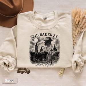 May include: Cream-coloured sweatshirt with a black and white graphic depicting 221B Baker St. in London, England, and a silhouette of a detective smoking a pipe. The text "EST. 1892" is above the detective. The sweatshirt has a crew neck.
