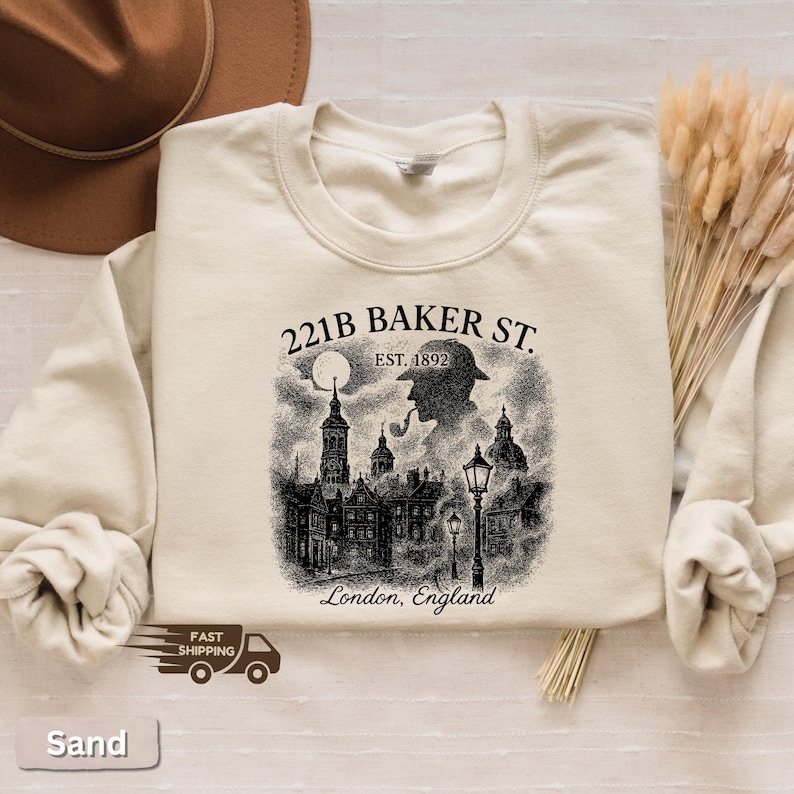 May include: Cream-coloured sweatshirt with a black and white graphic depicting 221B Baker St. in London, England, and a silhouette of a detective smoking a pipe. The text "EST. 1892" is above the detective. The sweatshirt has a crew neck.