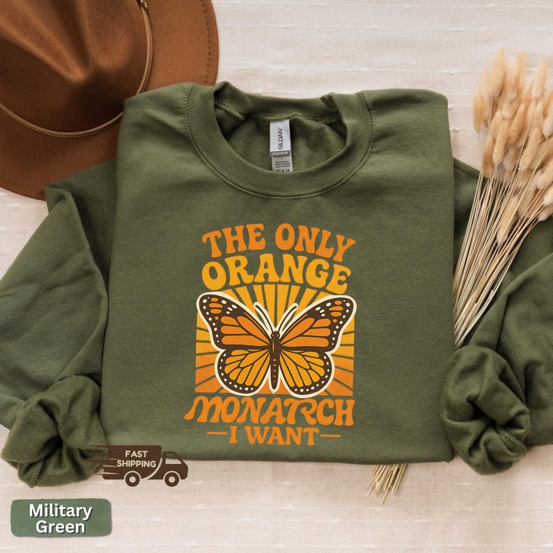 Op de afbeelding: Militairgroen sweatshirt met de tekst "THE ONLY ORANGE MONARCH I WANT" en een vlindergrafiek in oranje en bruin. Het sweatshirt is op een lichtgekleurd oppervlak gevouwen.
