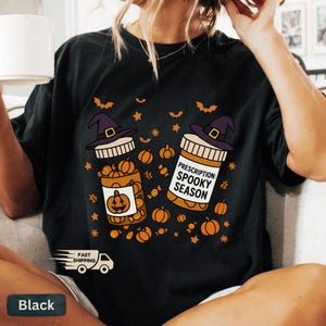 Op de afbeelding: Zwart T-shirt met een Halloween-thema. Het ontwerp toont twee pillenflessen met heksenhoeden, gevuld met pompoenen. E&eacute;n fles heeft een pompoenlabel, de andere luidt "Prescription: Spooky Season". Oranje vleermuizen en sterren maken het ontwerp compleet.