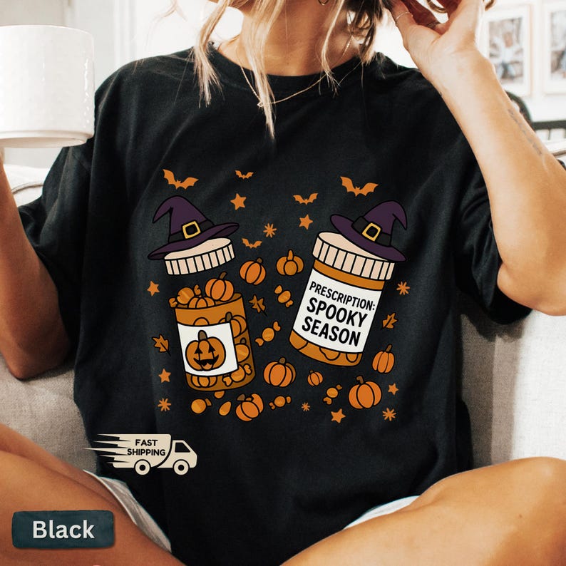 Op de afbeelding: Zwart T-shirt met een Halloween-thema. Het ontwerp toont twee pillenflessen met heksenhoeden, gevuld met pompoenen. E&eacute;n fles heeft een pompoenlabel, de andere luidt "Prescription: Spooky Season". Oranje vleermuizen en sterren maken het ontwerp compleet.