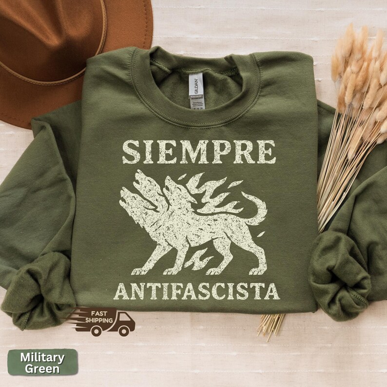 May include: Military green sweatshirt with the words "SIEMPRE ANTIFASCISTA" in white block letters. A three-headed wolf graphic is centered below the text. The sweatshirt is folded, with a brown hat and dried plants in the background.
