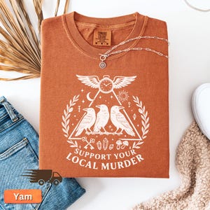 May include: A rust-colored t-shirt with a white graphic featuring three crows inside a triangle, a clock, and the text "SUPPORT YOUR LOCAL MURDER." A silver necklace with a pendant is draped over the shirt. The shirt is folded on a white surface.