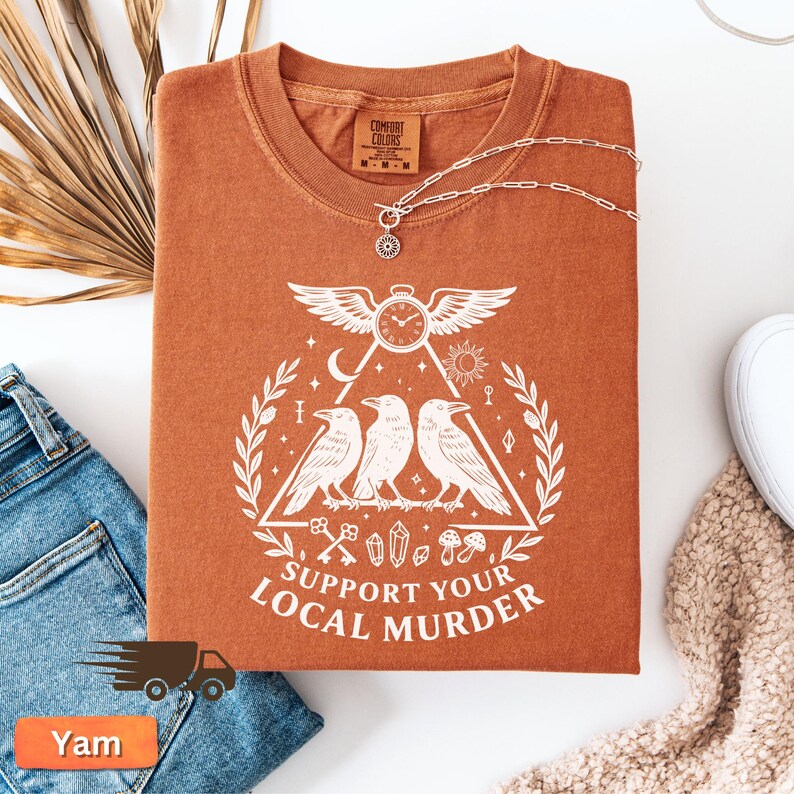 May include: A rust-colored t-shirt with a white graphic featuring three crows inside a triangle, a clock, and the text "SUPPORT YOUR LOCAL MURDER." A silver necklace with a pendant is draped over the shirt. The shirt is folded on a white surface.