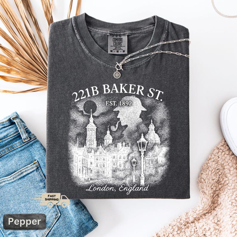 May include: Charcoal grey t-shirt featuring a graphic of 221B Baker St., London, England. The design includes a detective silhouette, buildings, and the text "EST. 1892". A silver necklace with a pendant is on the shirt.