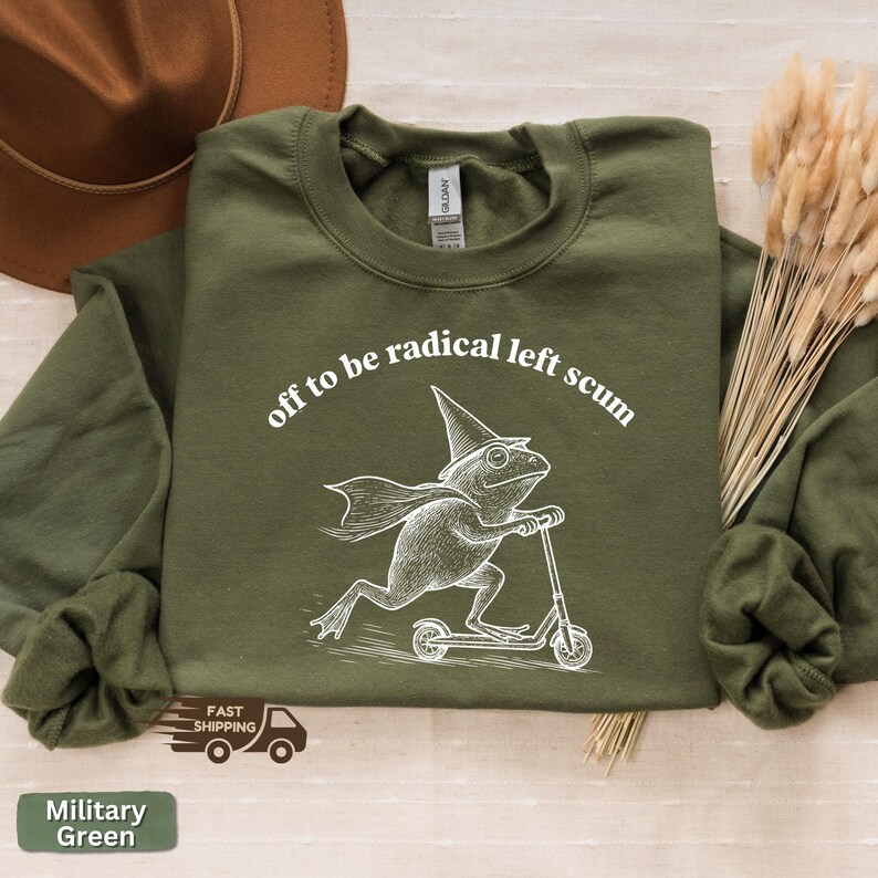 Pode incluir: Camisola verde militar com o texto "off to be radical left scum" e uma ilustra&ccedil;&atilde;o branca de um sapo numa scooter. A camisola tem gola redonda e mangas compridas.