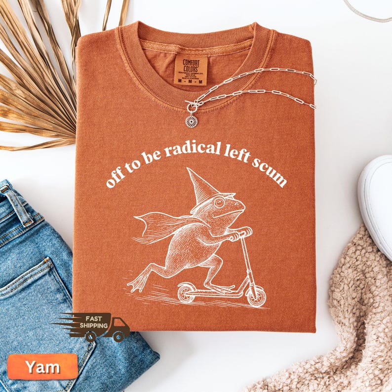 Pode incluir: Uma camiseta cor de ferrugem com o texto "off to be radical left scum" em arco. Uma ilustra&ccedil;&atilde;o branca de um sapo usando um chap&eacute;u de mago e capa em uma scooter est&aacute; abaixo. Um colar de prata est&aacute; sobre a camiseta.