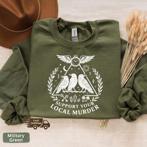 May include: Military green sweatshirt with a white graphic featuring three crows, a clock, and the text "SUPPORT YOUR LOCAL MURDER." The sweatshirt is made of a soft material and has a crew neck.