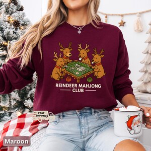 Puede incluir: Sudadera granate con un gr&aacute;fico de cuatro renos jugando al mahjong. El texto "REINDEER MAHJONG CLUB" est&aacute; impreso debajo de la ilustraci&oacute;n. La sudadera es de color granate liso.