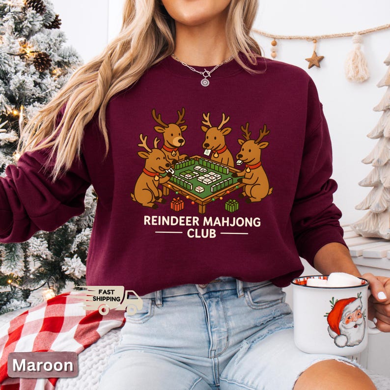 Puede incluir: Sudadera granate con un gr&aacute;fico de cuatro renos jugando al mahjong. El texto "REINDEER MAHJONG CLUB" est&aacute; impreso debajo de la ilustraci&oacute;n. La sudadera es de color granate liso.