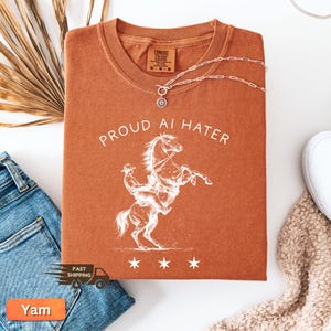 May include: A rust-colored t-shirt with the text "PROUD AI HATER" and a white graphic of a cowboy riding a rearing horse. A silver necklace with a sunburst pendant rests on the shirt. The shirt is folded, with blue jeans and a white shoe visible.