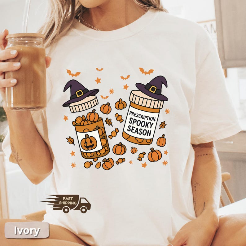 Op de afbeelding: Ivoor T-shirt met een Halloween-thema. Het ontwerp bevat twee pillenflessen met heksenhoeden, gevuld met pompoenen en snoep. De tekst "Prescription: Spooky Season" staat op een fles, met vleermuizen en sterren.